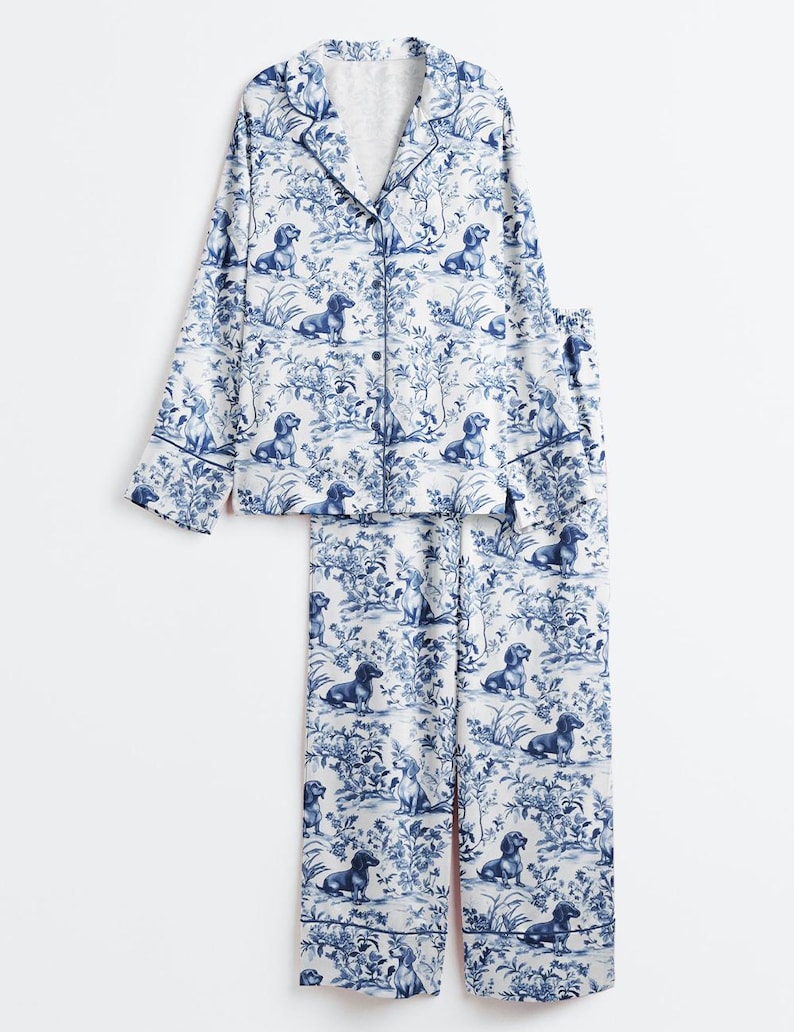 Puede incluir: Conjunto de pijama de dos piezas con un estampado de toile de Jouy azul y blanco con perros salchicha y elementos florales. La parte superior de manga larga tiene botones y los pantalones son de corte holgado.
