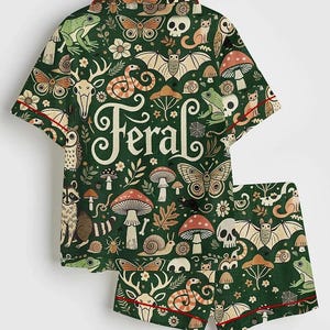 Op de afbeelding: Groene pyjamaset met een grillig patroon van bosdieren, paddenstoelen en het woord "Feral". De shirt met korte mouwen en de shorts hebben een rode bies. Het ontwerp bevat vleermuizen, slangen en herten.