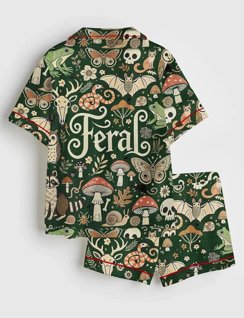 Op de afbeelding: Groene pyjamaset met een grillig patroon van bosdieren, paddenstoelen en het woord "Feral". De shirt met korte mouwen en de shorts hebben een rode bies. Het ontwerp bevat vleermuizen, slangen en herten.