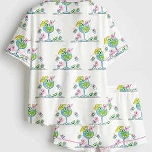 Puede incluir: Conjunto de pijama blanco con un estampado repetido de c&oacute;cteles azules con sombrillas amarillas y detalles en rosa. El conjunto incluye una camisa de manga corta con botones y pantalones cortos a juego con ribete rosa.
