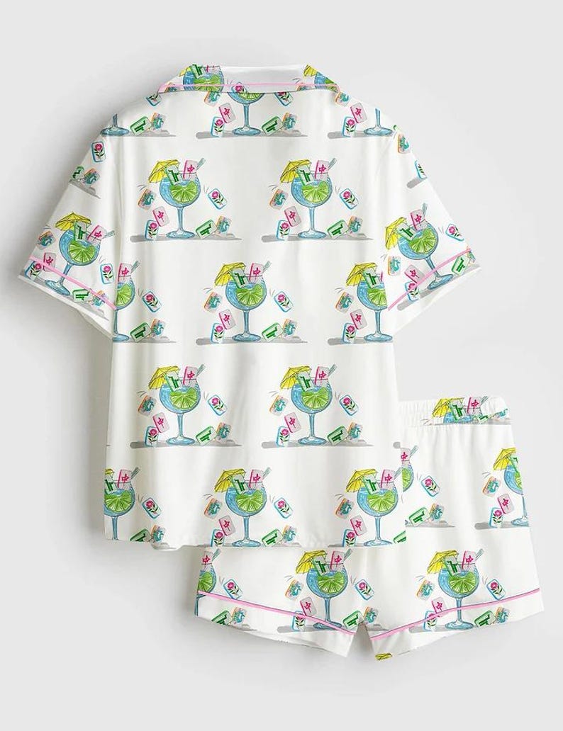 Puede incluir: Conjunto de pijama blanco con un estampado repetido de c&oacute;cteles azules con sombrillas amarillas y detalles en rosa. El conjunto incluye una camisa de manga corta con botones y pantalones cortos a juego con ribete rosa.