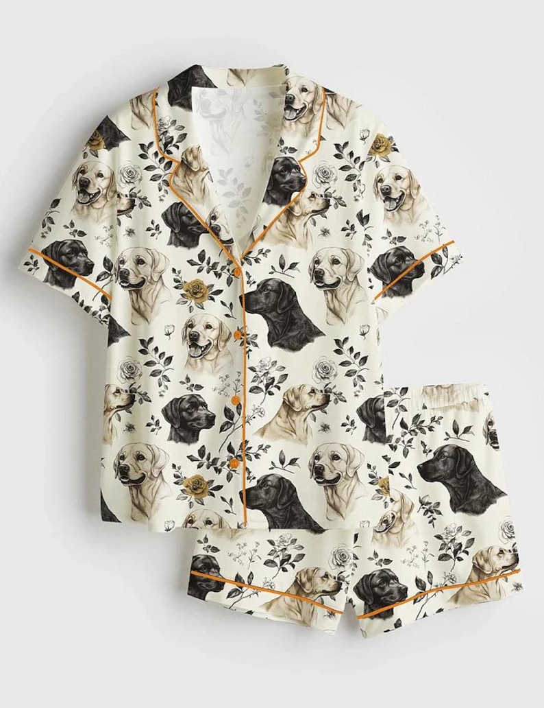 May include: Cream-colored pajama set featuring a repeating pattern of Labrador retrievers and floral accents. The short-sleeved top and shorts have orange trim. The pajama set includes a button-down shirt and matching shorts.