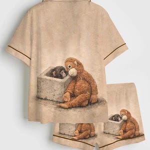 May include: Tan pajama set featuring a whimsical illustration of two orangutans. The short-sleeved top and shorts have brown trim. One orangutan sits next to a smaller one in a stone box. The set is made of a soft material.