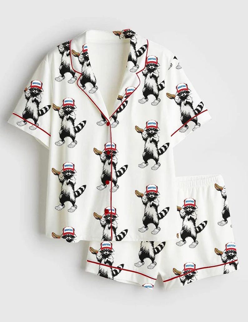 Op de afbeelding: Witte pyjamaset met een herhaald patroon van cartoon wasberen met hoeden met het woord "America" en een snack. De set bevat een shirt met korte mouwen en een korte broek, beide afgezet met rode biezen.