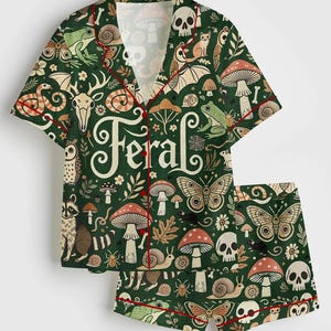 Op de afbeelding: Groene pyjamaset met een grillig bospatroon met paddenstoelen, schedels, vlinders en bosdieren. De set bevat een overhemd met knopen en een korte broek, beide afgezet met rode biezen. Het woord "Feral" staat op het shirt.