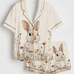 May include: Cream-colored pajama set featuring a watercolor illustration of a white rabbit amidst wildflowers. The short-sleeved top and shorts have brown trim. The bunny design is repeated on both pieces, creating a cohesive sleepwear set.