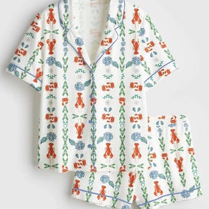 May include: White pajama set with a lobster and floral print. The short-sleeved top and shorts feature a repeating pattern of red lobsters, blue flowers, and green vines, outlined with blue trim.
