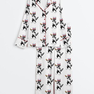Op de afbeelding: Witte pyjamaset met een herhalend patroon van wasberen met honkbalpetten en knuppels. De top met lange mouwen heeft rode biezen en knopen. De pyjamabroek heeft ook rode afwerking. Een leuke en speelse nachtkledingset.