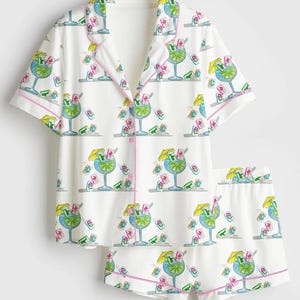 Puede incluir: Conjunto de pijama blanco con estampado de c&oacute;cteles. La parte superior de manga corta y los pantalones cortos presentan un patr&oacute;n repetido de c&oacute;cteles azules con rodajas de lima, sombrillas amarillas y detalles en rosa. Los bordes est&aacute;n ribeteados con ribete rosa.