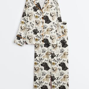 May include: A two-piece pajama set featuring a repeating pattern of black and golden Labrador retrievers and floral accents on a cream background. The set includes a button-down top with orange trim and matching pants.