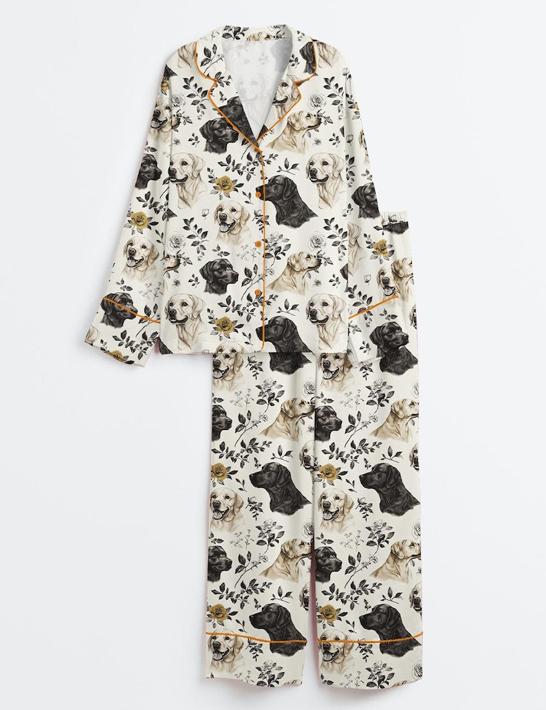 May include: A two-piece pajama set featuring a repeating pattern of black and golden Labrador retrievers and floral accents on a cream background. The set includes a button-down top with orange trim and matching pants.