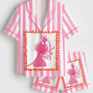 May include: Pink and white striped pajama set. The short-sleeved top and shorts feature a peacock graphic with the text "bird bam cheers" inside a decorative frame. The set has pink trim.