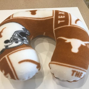 May include: A brown and white travel pillow with a University of Texas Longhorns pattern. The pillow features the Longhorn logo, a football helmet, and the word "TEX".