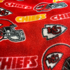 K&ouml;nnte beinhalten: Roter Fleece-Stoff mit einem sich wiederholenden Muster des Logos der Kansas City Chiefs, einschlie&szlig;lich des Teamnamens "Chiefs" und des Pfeilspitzen-Logos mit "KC" darin.
