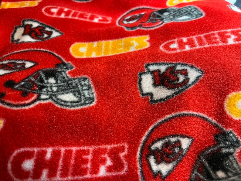 K&ouml;nnte beinhalten: Roter Fleece-Stoff mit einem sich wiederholenden Muster des Logos der Kansas City Chiefs, einschlie&szlig;lich des Teamnamens "Chiefs" und des Pfeilspitzen-Logos mit "KC" darin.