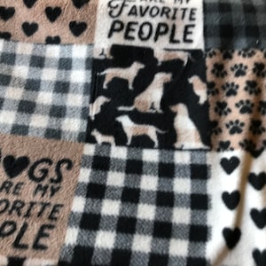 May include: A patchwork fleece blanket featuring various patterns and dog-themed designs. The blanket includes squares with black and white buffalo check, dog silhouettes, paw prints, and hearts. Text reads "Dogs are my favorite people."