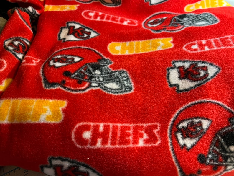 K&ouml;nnte beinhalten: Roter Fleece-Stoff mit einem sich wiederholenden Muster aus wei&szlig;em "Chiefs"-Text, einem wei&szlig;en Pfeil mit "K.C." darin und einem wei&szlig;en Footballhelm mit schwarzen Details.