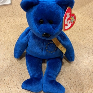 May include: A plush blue teddy bear with black eyes and a gold ribbon. The bear has a red heart-shaped tag with the word "Beanie" on it. The bear is approximately 8 inches tall.
