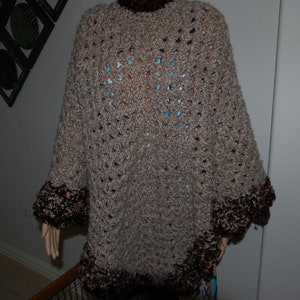 May include: A handmade, open-weave poncho in shades of brown and tan. The poncho features a high collar and a decorative trim of dark brown and reddish-brown yarn along the bottom edge and sleeve openings. The garment is displayed on a mannequin.