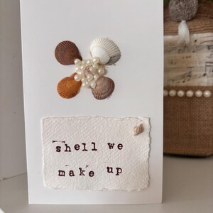 Puede incluir: Una tarjeta de felicitación blanca con un diseño de conchas marinas. La tarjeta presenta un grupo de conchas marinas en tonos marrón, naranja y blanco, acentuadas con pequeñas perlas. El texto "shell we make up" está impreso en un parche de papel texturizado.