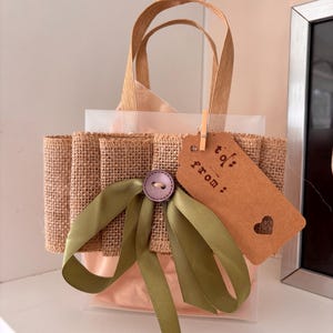 Può includere: Un piccolo sacchetto regalo con rifiniture in juta e nastro verde oliva. Un'etichetta marrone con le parole "to: from:" e un cuore è attaccata con una piccola clip di legno. La borsa ha un bottone e un manico.
