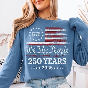 May include: A long-sleeve, blue shirt featuring a distressed American flag design. The flag includes the text "1776" in a circle of stars, and the words "We The People" and "250 YEARS 2026" in white.