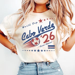 May include: A cream-colored short-sleeve t-shirt featuring a graphic with the text World Cup Cabo Verde '26 Soccer National. The design includes a soccer ball, a red star, blue stars, and the Cabo Verde flag in red, white, and blue colors.