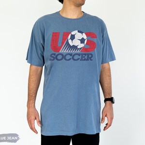 May include: A blue jean colored short-sleeve t-shirt featuring a graphic print with the text US SOCCER in red and dark blue. The design includes a white and dark blue soccer ball illustration with motion lines, all rendered in a distressed texture.