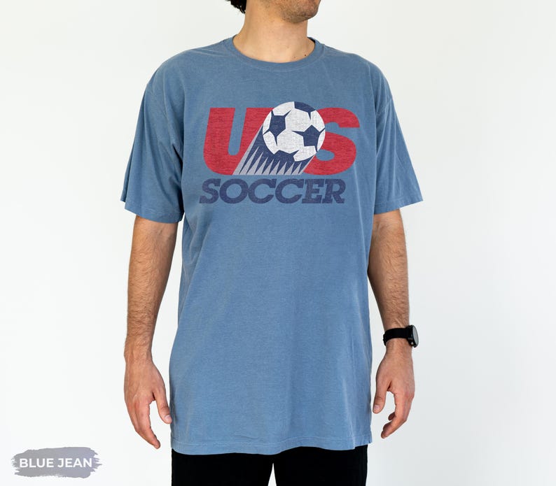 May include: A blue jean colored short-sleeve t-shirt featuring a graphic print with the text US SOCCER in red and dark blue. The design includes a white and dark blue soccer ball illustration with motion lines, all rendered in a distressed texture.