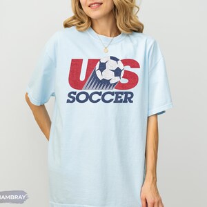 May include: A light blue short-sleeve t-shirt featuring a graphic design with the red text US, a black and white soccer ball, and the blue text SOCCER. The graphic has a distressed texture. The word CHAMBRAY appears in a small gray box at the bottom.