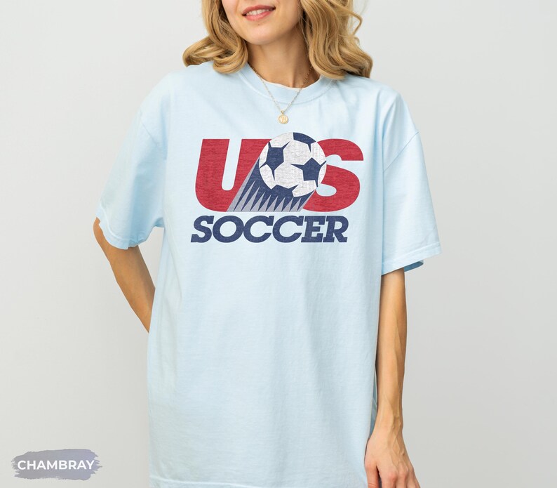 May include: A light blue short-sleeve t-shirt featuring a graphic design with the red text US, a black and white soccer ball, and the blue text SOCCER. The graphic has a distressed texture. The word CHAMBRAY appears in a small gray box at the bottom.