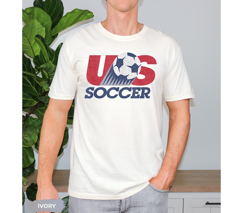 May include: An ivory short-sleeve t-shirt featuring a graphic design with the red letters US, a black and white soccer ball with a motion trail, and the word SOCCER printed in dark blue capital letters below the graphic on the chest.
