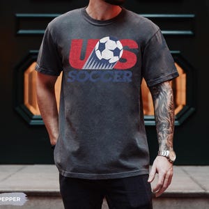 May include: A charcoal gray short-sleeve t-shirt featuring a distressed graphic print. The design displays the red letters US with a white and black soccer ball in the center, and the word SOCCER in blue text underneath the ball graphic.