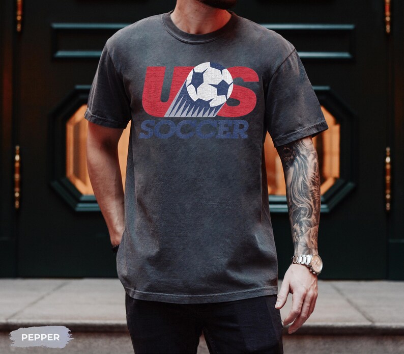 May include: A charcoal gray short-sleeve t-shirt featuring a distressed graphic print. The design displays the red letters US with a white and black soccer ball in the center, and the word SOCCER in blue text underneath the ball graphic.