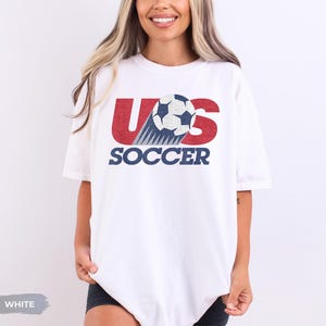 May include: A white short-sleeved t-shirt featuring a graphic design with the red letters US, a black and white soccer ball replacing the letter O, and the word SOCCER printed in dark blue capital letters underneath the graphic.