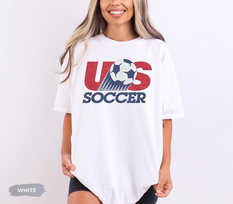 May include: A white short-sleeved t-shirt featuring a graphic design with the red letters US, a black and white soccer ball replacing the letter O, and the word SOCCER printed in dark blue capital letters underneath the graphic.