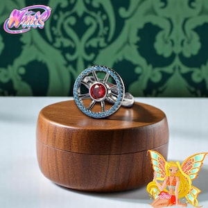 Könnte beinhalten: Ein silberner Ring mit kreisförmigem Design, einem zentralen roten Stein und einem Kranz aus kleinen blauen Steinen. Der Ring wird auf einem Holzständer präsentiert. Ein Cartoon-Feenbild befindet sich in der Ecke.