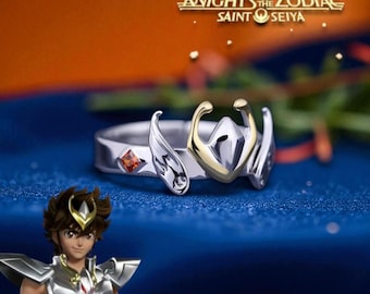 May include: A silver ring with gold accents and a small orange gemstone. The ring features a stylized design with the text "Knights of the Zodiac Saint Seiya" in gold lettering. The ring is set against a blue and orange background.