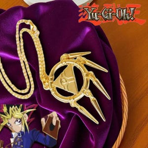 Pode incluir: Um colar com pingente dourado com um design estilizado, apresentando um triângulo central e três pontas salientes. O colar repousa sobre um tecido de veludo roxo, com o logotipo "Yu-Gi-Oh!" ao fundo.