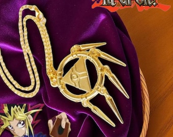 May include: A gold-colored pendant necklace featuring a stylized design with a central triangle and three outward-pointing prongs. The necklace rests on a purple velvet surface, with the "Yu-Gi-Oh!" logo visible in the background.