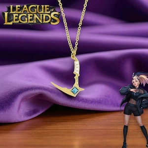 以下が含まれることがあります： 青い宝石と白いアクセントが特徴の、金色のペンダントネックレス。スタイリッシュな鎌のデザインです。ネックレスは、紫色の背景に金のチェーンで吊り下げられています。「League of Legends」のロゴが見えます。