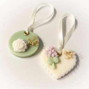 May include: Two decorative wax ornaments with white ribbon hangers. One is a light green circle with a white rose and dried flowers. The other is a heart shape with a lavender and green floral design and dried flowers. Both have gold eyelets.