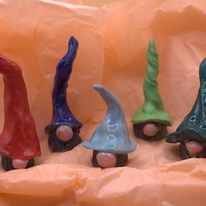 May include: Five ceramic gnome figurines with colourful hats in red, blue, green, and teal. Each gnome has a round, pink nose and a dark, textured base. The gnomes are arranged on a peach-coloured, textured surface.