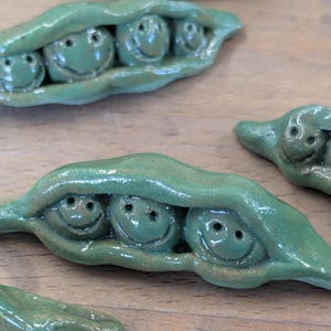 May include: Handmade ceramic pea pods, each with three smiling peas. The pods are a glossy green with a textured surface, and the peas have simple, happy faces. The whimsical design is set against a wooden background.