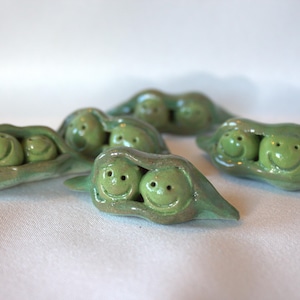 May include: A collection of four small, green ceramic pea pods, each featuring two smiling faces carved into the peas nestled inside the open pod. The glazed finish gives the pieces a smooth, reflective appearance with varied shades of green.