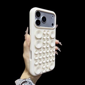 Puede incluir: Una funda de teléfono blanca con un diseño único que presenta una serie de protuberancias circulares en relieve. La funda tiene un acabado liso y un recorte para la cámara. El fondo es negro.