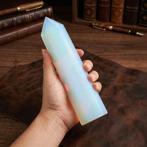 May include: A translucent, opalescent crystal obelisk with a pointed top. The crystal is held in a hand, showcasing its smooth, polished surface. The obelisk is approximately 6 inches tall, with a soft, glowing interior.