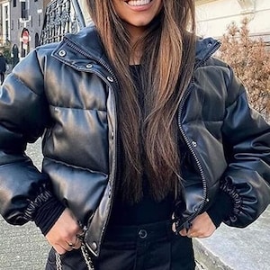 Puede incluir: Chaqueta acolchada negra con cuello alzado, cierres a presión y cremallera. La chaqueta tiene mangas largas con puños acanalados. La modelo lleva un top negro y pantalones negros. La chaqueta es de material brillante de cuero sintético.