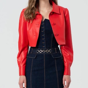 May include: A bright red, cropped leather jacket with long sleeves and a collared neckline. The jacket features button closures and is paired with a dark denim dress. The outfit is completed with a silver chain belt.
