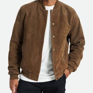 Puede incluir: Chaqueta bomber de ante marrón con cuello, puños y dobladillo de canalé. La chaqueta tiene cierre de botones y dos bolsillos laterales. La chaqueta es de color marrón cálido y tiene un diseño clásico.